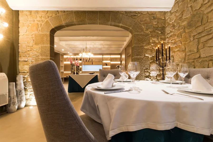 Mesa redonda puesta con mantel blanco y copas, en un comedor de muros de piedra con un arco al fondo