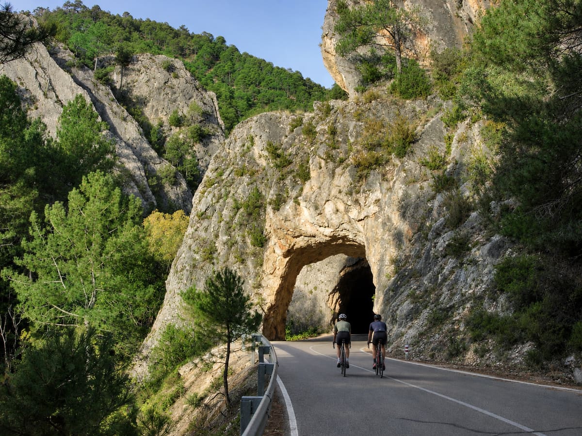 Dos ciclistas de carretera ante un túnel excavado en la roca, entre paredes calizas y pinares de la Serranía de Cuenca