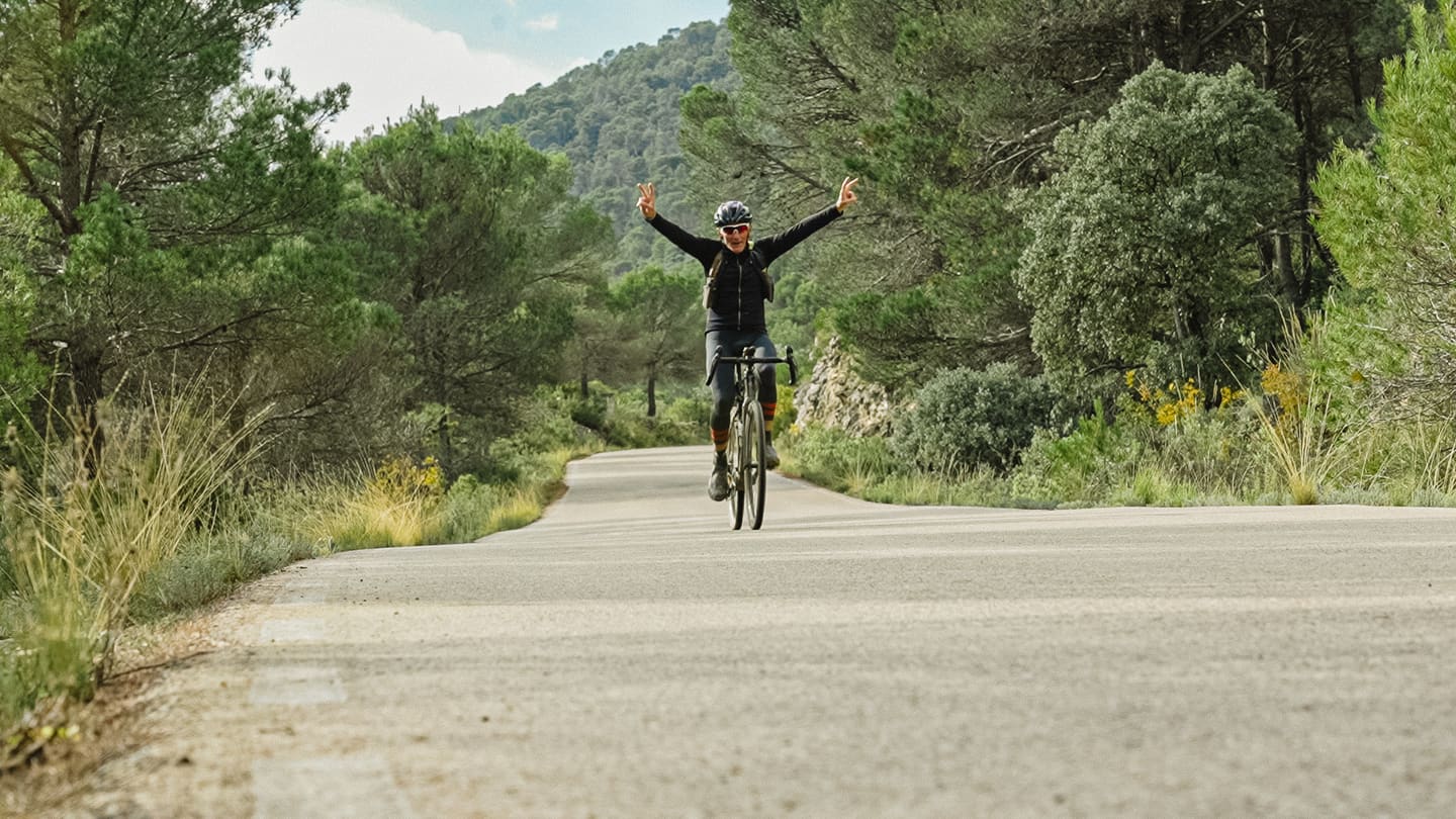 Ciclista de carretera con los brazos en alto, pedaleando por una carretera estrecha entre pinares