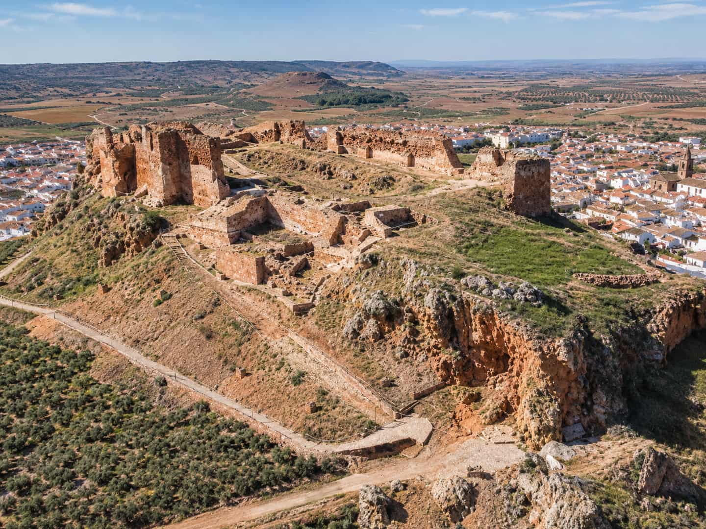 Vista aérea de las ruinas de un castillo sobre un cerro, con un pueblo blanco y campos de olivar al fondo