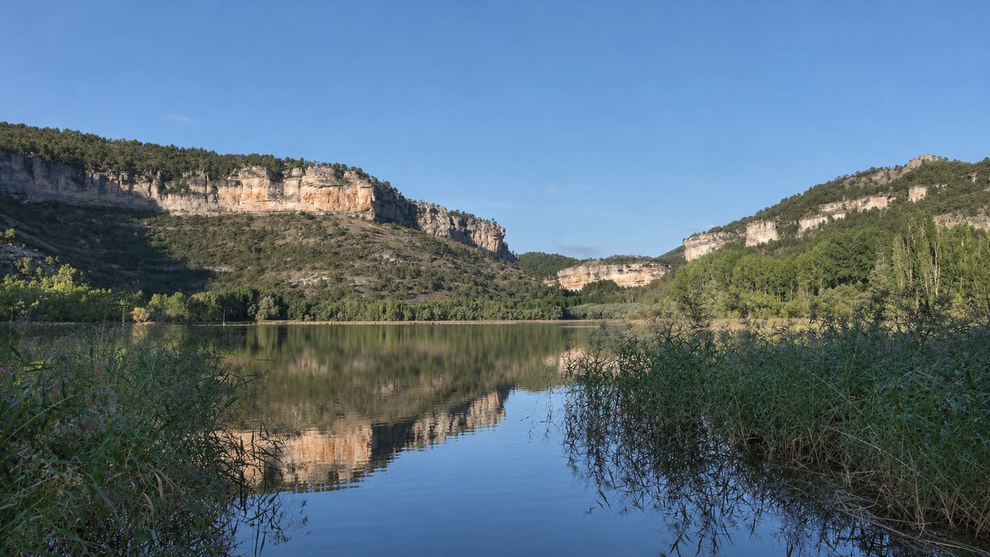 Cortados de roca caliza y pinares reflejados en las aguas tranquilas de una laguna, entre juncos