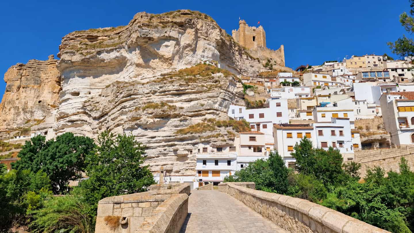 Pueblo de casas blancas escalonado bajo una gran peña caliza coronada por un castillo, con un puente de piedra en primer plano
